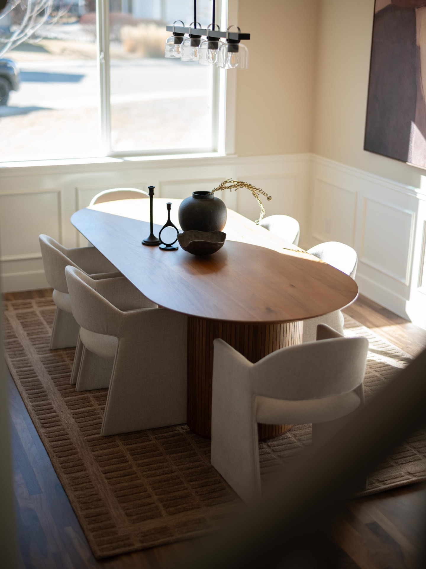 Oval walnut dining table on a woven rug, styled with a matte black vase and candlesticks beneath a linear glass chandelier