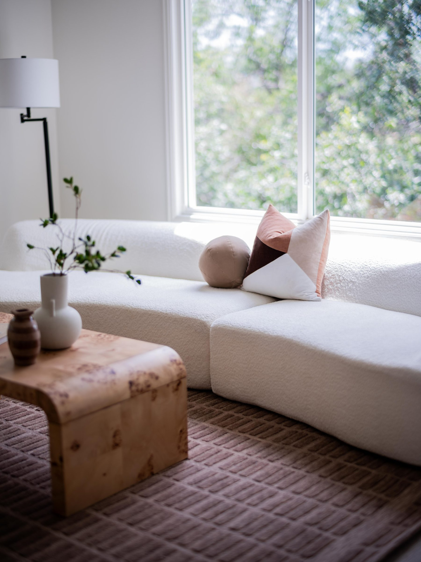 Bouclé sofa beneath a leafy window with blush pillows and a burl wood coffee table on a plum grid rug