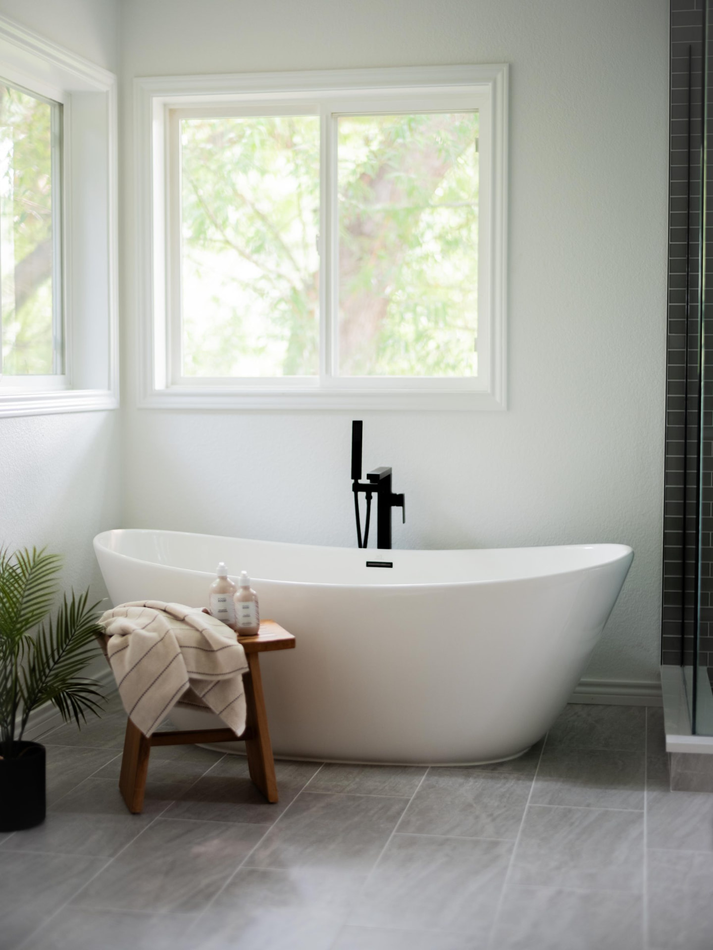 Freestanding soaking tub with a matte black filler, teak bench and greenery framed by a bright window